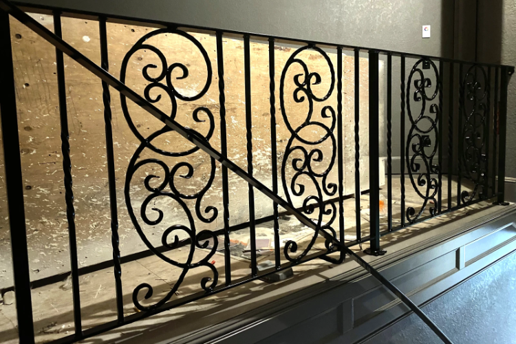 A custom-handcrafted interior mezzanine railing featuring ornate black wrought iron scrollwork panels set between vertical balusters.